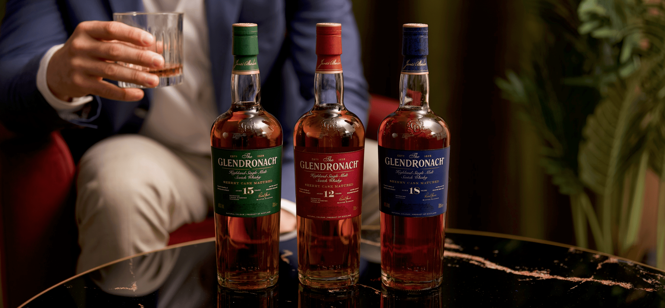 An image of three bottles of The Glendronach Whisky on a table with a man holding a glass in the background.