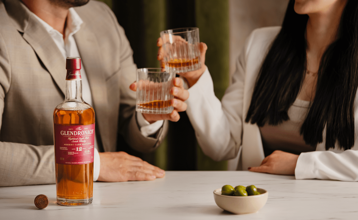 An image of a bottle of The Glendronach Scottish Whisky in the forefront with a man and women in the background toasting their glasses at a table.